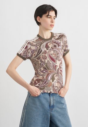 Giovane in piedi con le mani nelle tasche, indossa una camicia a maniche corte con motivo paisley e jeans blu larghi su sfondo neutro.