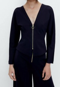 Top lungo nero a maniche lunghe con scollatura a V profonda, dotato di una zip frontale con un tirante circolare, design aderente e texture liscia.