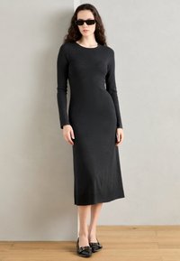 Black, long-sleeve dress made of a smooth, stretchy fabric with a fitted silhouette that falls to mid-calf, paired with flat, embellished shoes.