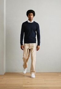 Navy sweater, collared white shirt, beige trousers, and white sneakers. Standing pose, smooth surfaces, hardwood floor, neutral background.