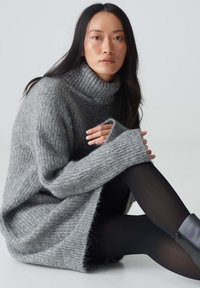 Grauer Strickpullover mit hohem Rippkragen und weiten Ärmeln, getragen über schwarzen Leggings und Stiefeletten, mit lockerer Passform und weicher Textur.
