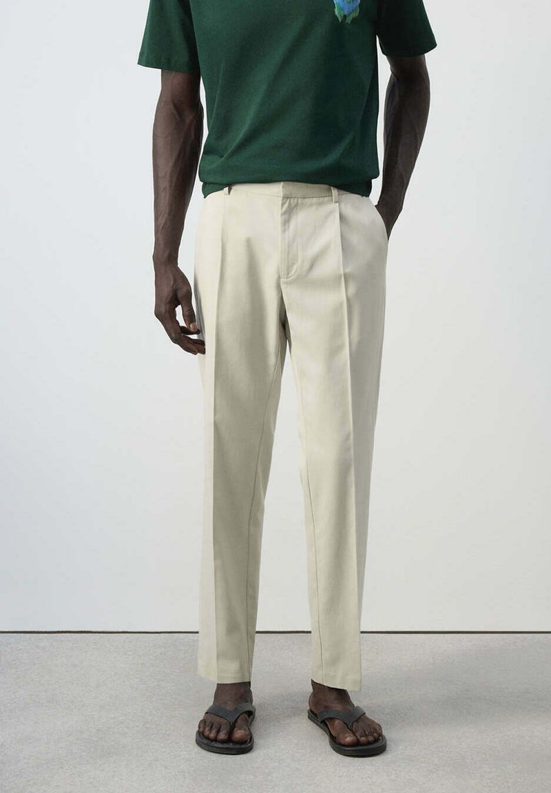 Homme portant un pantalon plissé beige, une chemise à manches courtes vert foncé et des sandales noires, debout sur un sol gris.