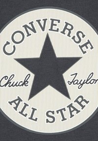 Juodos ir kreminės spalvos apskritas logotipas su ryškia juoda žvaigžde, kurioje parašyta „CONVERSE“ ir „ALL STAR“ ryškiu šriftu, o „Chuck Taylor“ – kursyvu.