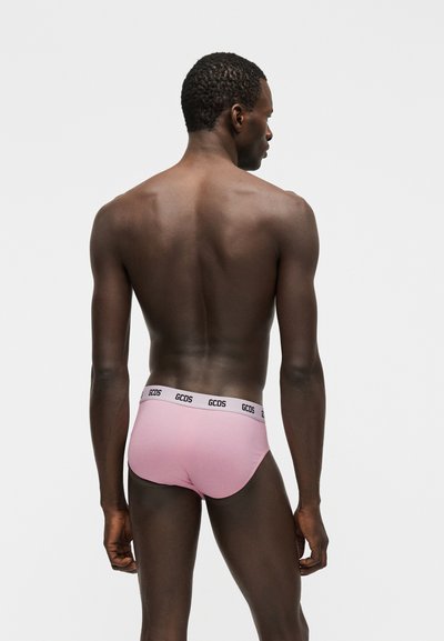 Άνδρας με σκούρο δέρμα, γυρισμένος πλάτη, φοράει ροζ εσώρουχα με λευκή μέση που φέρει τη μάρκα «GCDS», στέκεται μπροστά σε απλό λευκό φόντο.