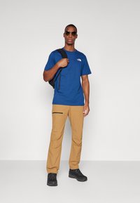 Zila īsām piedurknēm T-krekls ar mazu baltu logo, gaiši brūnas bikses ar sānu rāvējslēdzēja kabatu un melniem pārgājienu apaviem, ko valkā ar saulesbrillēm un mugursomu.