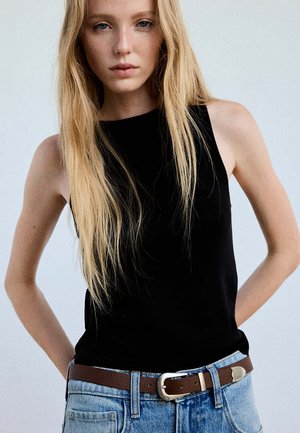 Jeune femme aux longs cheveux blonds portant un haut noir sans manches, un jean bleu clair et une ceinture marron, debout devant un fond uni.