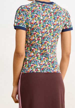 Frau trägt ein kurzärmeliges Hemd mit Blumenmuster und marineblauem Besatz, kombiniert mit einem braunen Rock mit zwei rosa vertikalen Streifen.