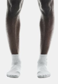 Calcetines tobilleros blancos con puños acanalados y una textura suave, llevados en piernas bien definidas y musculosas sobre un fondo blanco.
