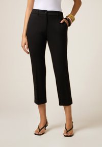 Femme portant un pantalon noir cropped avec des ourlets retroussés, des sandales à talons noires, un haut blanc et plusieurs bracelets colorés sur un poignet.