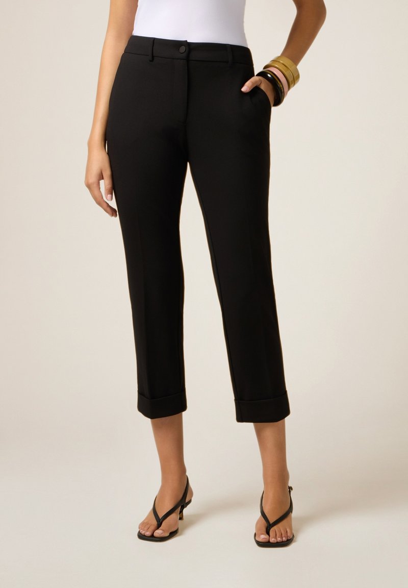 Femme portant un pantalon noir cropped avec des ourlets retroussés, des sandales à talons noires, un haut blanc et plusieurs bracelets colorés sur un poignet.