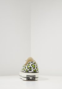 Białe sportowe buty bez zapięcia z zielono-czarnym wzorem w panterę, z gumową podeszwą i czarnym logo na pięcie.