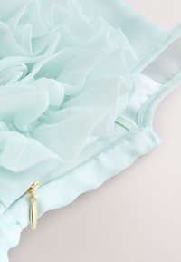 Détail d'une robe bleu clair avec tissu transparent superposé, fermeture éclair visible avec tirette dorée, et bretelle cousue sur un fond pâle.