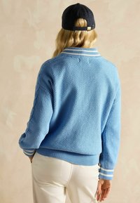 Pulover tricotat de culoare albastru deschis, cu un model texturat, manșete și guler striate, prezentând dungi albe. Purtați cu pantaloni de culoare crem.