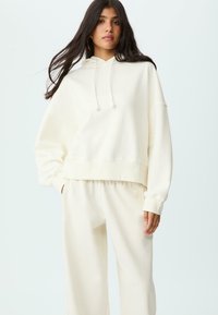 Ung kvinna med långt mörkt hår, iförd en överdimensionerad krämfärgad hoodie och matchande sweatpants, som står mot en enfärgad ljus bakgrund.