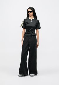 Woman with long black hair wearing black sunglasses, a black Adidas shirt with yellow stripes, wide-leg black pants, and gray sneakers.