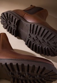 Hnedé kožené členkové topánky s elastickými bočnými panelmi, vybavené čiernou textúrovanou gumovou podrážkou s hlbokým dezénom pre lepšiu trakciu.