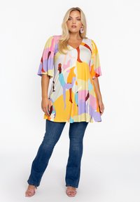 Tunique colorée à manches courtes avec un décolleté en V, ornée de formes abstraites en jaune, rose, violet et bleu. Associée à un jean foncé.
