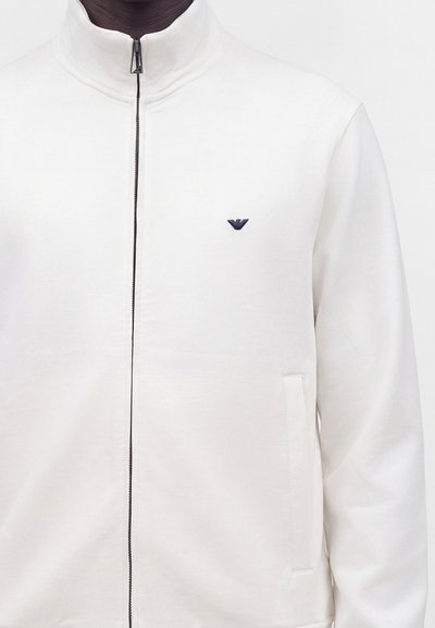 Homme portant une veste blanche zippée unie avec un petit logo sombre sur le côté gauche de la poitrine et des poches latérales visibles.