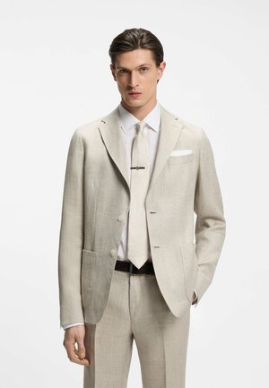 Homme portant un costume en lin beige clair avec une cravate assortie, une chemise blanche, une pince à cravate et une pochette, debout devant un fond blanc uni.