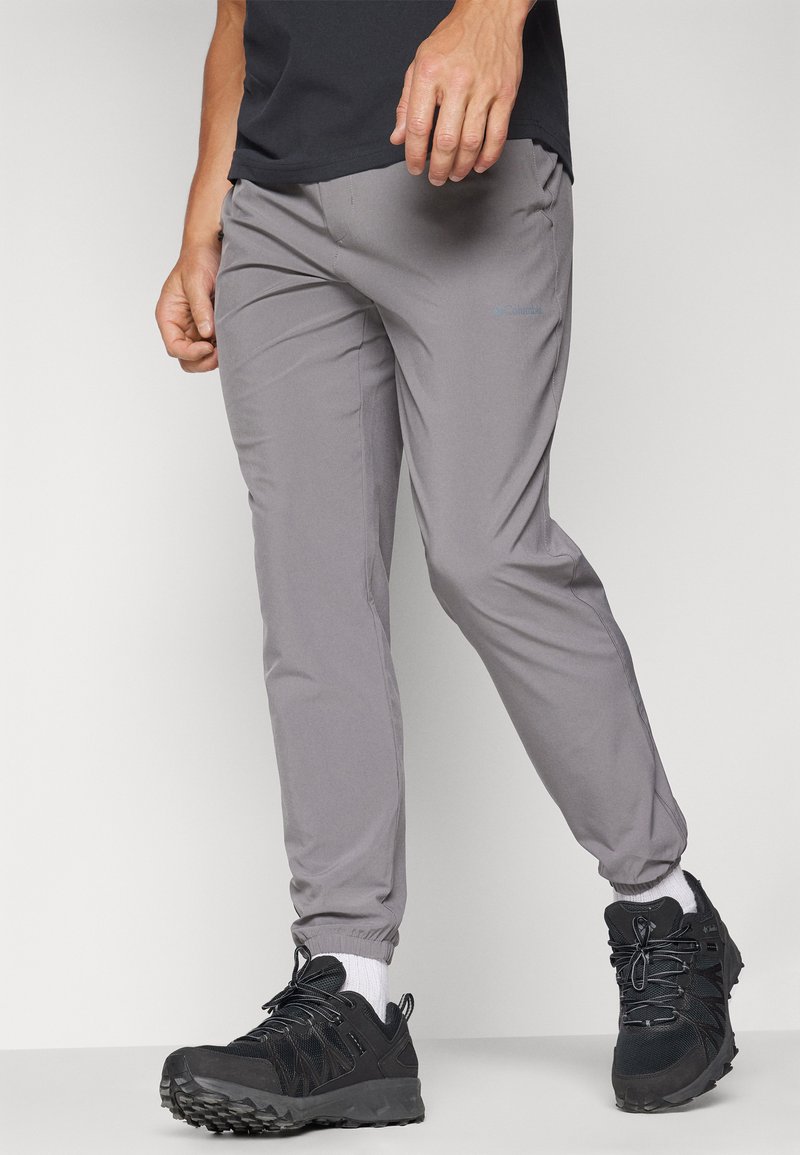 Mann in grauen, schmal zulaufenden Jogginghosen, schwarzen Sportschuhen, weißen Socken und einem schwarzen Shirt vor einem einfarbigen hellen Hintergrund.