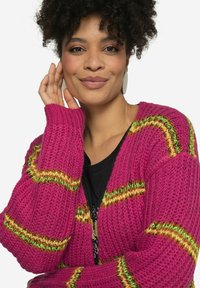 Roze gebreide cardigan met groene en gele strepen, losse pasvorm, geribbelde mouwen en een gestructureerde stof. Draagt over een zwarte top.