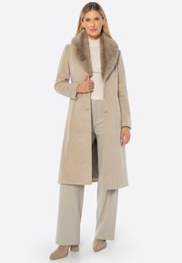 Beżowy wełniany płaszcz z dużym kołnierzem z sztucznego futra, o prostym kroju, zapinany na jeden rzęd guzików z przodu.