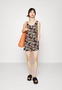 Macacão sem mangas floral com folhas verdes e flores pink sobre um fundo azul-marinho. Acompanhado de uma mala laranja e sandálias pretas.