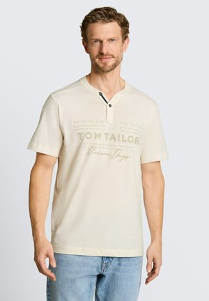 Mężczyzna o jasnobrązowych włosach, ubrany w kremową koszulkę z krótkim rękawem z napisem "Tom Tailor Riviera Days" i niebieskie dżinsy, stojący na jasnym tle.