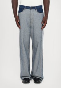 Homme portant un jean en denim à jambes larges bicolores avec des poches bleu foncé et des chaussures noires, se tenant devant un fond blanc uni.