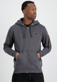 Felpa grigia con zip, dotata di tasca frontale, cappuccio con cordino, tasca laterale con zip e un patch con logo sul petto. Tessuto di texture liscia.