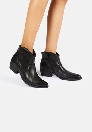 Jambes portant des bottines en cuir noir avec talons bas et carrés et des fermetures éclair latérales sur fond blanc.