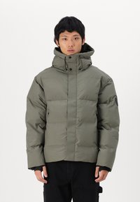 Puffer jacket in olive green, featuring a high collar, zippered front, and two side pockets. Soft texture with a padded design and black logo patch.