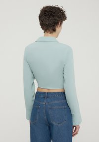 Blusa verde menta accorciata con maniche lunghe e colletto, indossata su jeans blu a vita alta. Tessuto liscio con un design aderente.