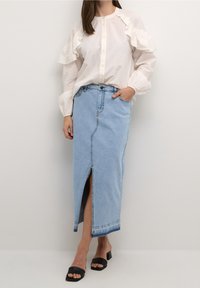 Blusa blanca con mangas acampanadas, combinada con una falda maxi de mezclilla azul claro con una abertura frontal. Sandalias de tacón cuadrado negras.