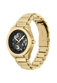 Złoty stalowy zegarek automatyczny ze czarnym szkieletem tarczy, złotymi wskazówkami i dwoma zielonymi subtarcza, na metalowej bransolecie.