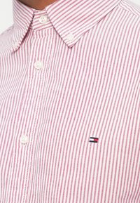 Rød og hvit stripete skjorte med knapp- ned krage, utstyrt med hvite knapper og en liten brodert logo i marineblått, rødt, og hvitt.