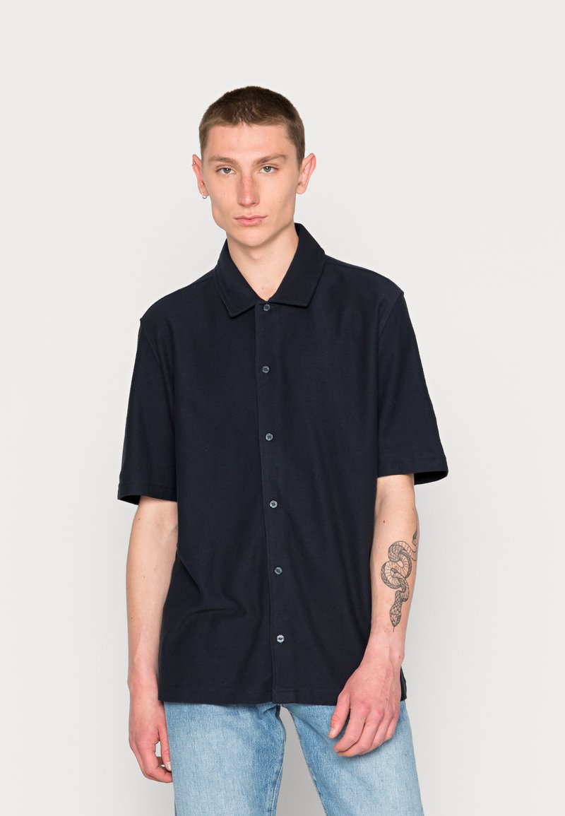 Camisa azul marinho de manga curta, com colarinho pontudo, feita de um tecido suave. Apresenta um corte descontraído e botões na parte da frente.
