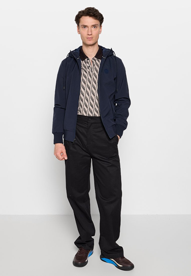 Giacca blu navy con cappuccio sopra una camicia a maniche corte a motivo, pantaloni neri e sneaker scure con dettagli blu. Outfit moderno e casual.