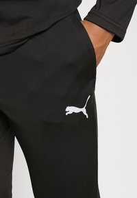 Juoksuhousut mustia urheiluvaatteita, valmistettu sileästä kankaasta, jossa on sivutaskut ja valkoinen Puma-logo vasemmassa reidessä.