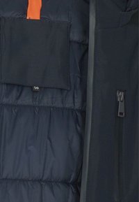 Primo piano dell'interno di una giacca imbottita blu scuro con una tasca sul petto e una cerniera nera sul lato destro.