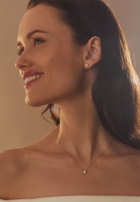 Gold necklace with a small diamond pendant. Model wears stud earrings, featuring a round design with inset diamonds, displaying smooth hair and neckline.