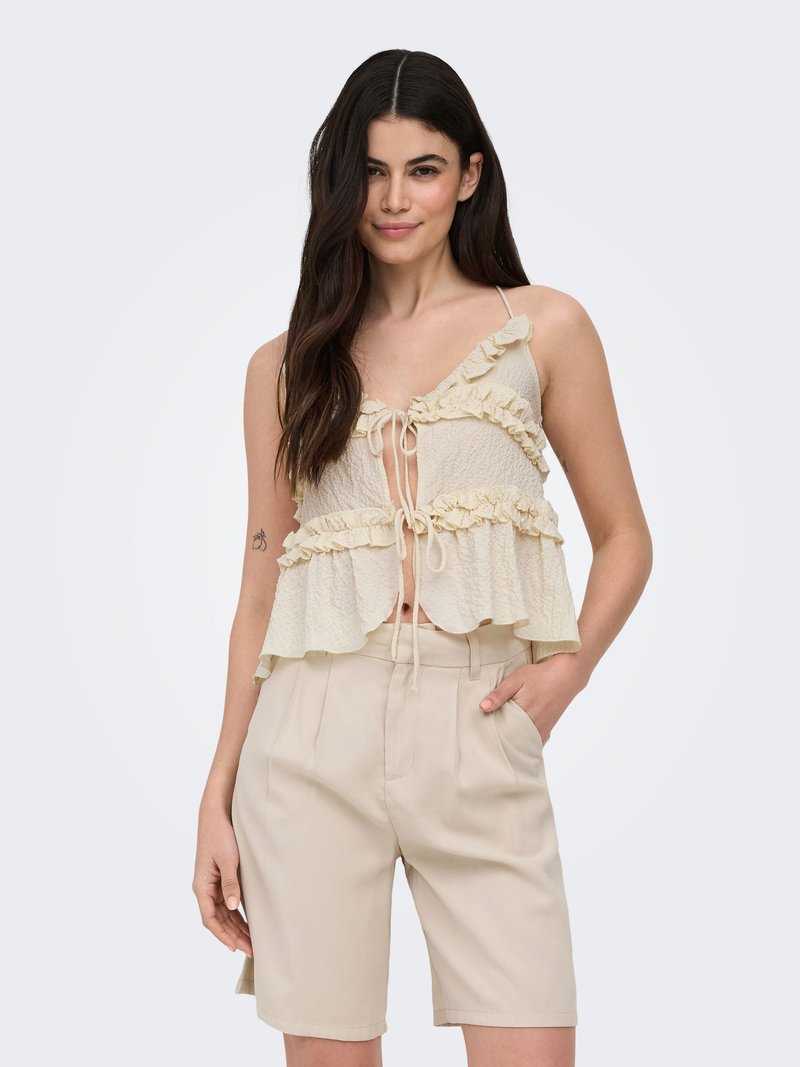 Femme aux longs cheveux foncés porte un crop top crème à volants avec des liens et un short beige clair jusqu’aux genoux, debout avec une main dans la poche.