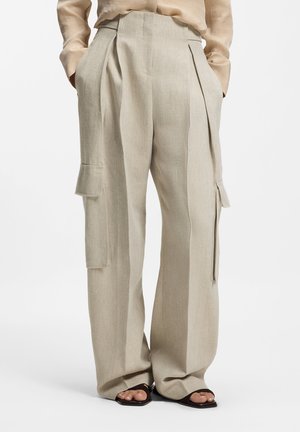 Beige Leinen-Cargohose mit weitem Bein, vorderen Falten und seitlichen Klappentaschen, kombiniert mit beige Oberteil und schwarzen Sandalen mit quadratischer Spitze.