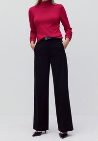 Enger Rollkragenpullover in leuchtendem Pink, kombiniert mit weit geschnittenen schwarzen Hosen. Schwarzer Gürtel mit dekorativer silberner Schnalle. Schwarze High Heels.