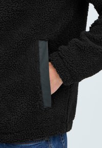 Giacca in pile nera con una superficie texturizzata e pelosa, dotata di una tasca laterale con una bordatura nera a contrasto. Mano nella tasca, jeans visibili sotto.