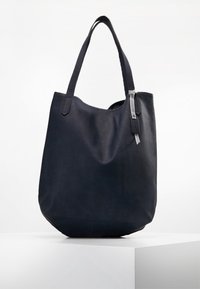 Svart tygpåse med texturerat material, dubbla axelremmar, mjuk, slapp form och en liten accentetikett fäst med ett band.