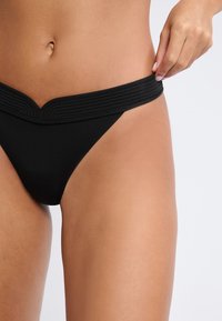 Bas de bikini noir avec une ceinture large en V texturée. Tissu lisse, couverture minimale, sans motifs ni éléments visibles.