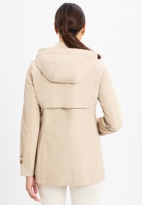 Beige wasserabweisende Jacke mit Kapuze, mit Reißverschluss und geknöpften Ärmeln, glatte Textur und mit einem Rückenbelüftung für zusätzlichen Bewegungsspielraum.