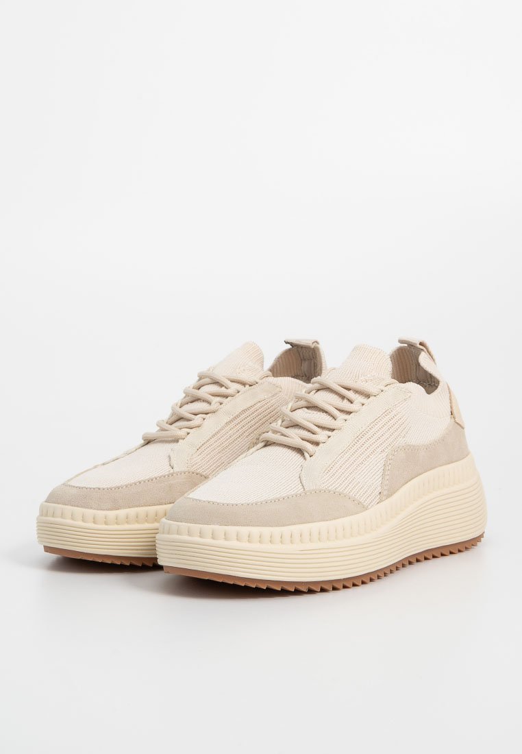 Beige Sneaker mit einem strukturierten Obermaterial aus Stoff und Wildlederakzenten, ausgestattet mit einer dicken, geriffelten Sohle und traditionellem Schnürdesign.