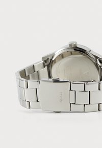 Reloj de metal con pulsera de acero inoxidable plateado, caja redonda plateada y un fondo liso y reflectante con detalles del logo grabados.